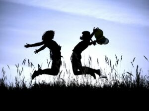 Silhouette of two children jumping joyfully in a field with balloons against a blue sky.